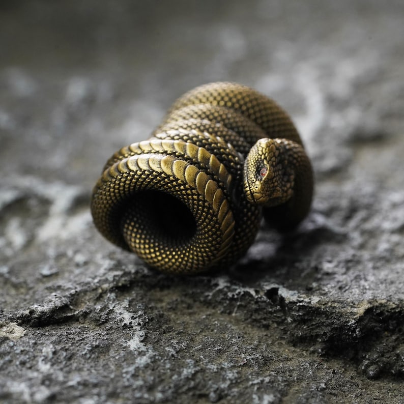 May include: A coiled brass snake sculpture with detailed scales and a red eye. The snake is positioned on a textured gray stone surface, creating a contrast between the metallic sheen and the rough stone.