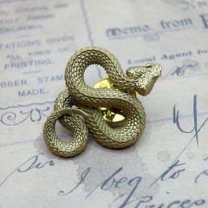 Könnte beinhalten: Ein goldfarbener Metall-Schlangen-Ansteckpin mit detaillierten Schuppen. Die Schlange ist aufgerollt, der Kopf ist erhoben. Der Pin hat eine goldfarbene Rückseite. Der Hintergrund ist ein Vintage-Papier mit handschriftlichem Text.