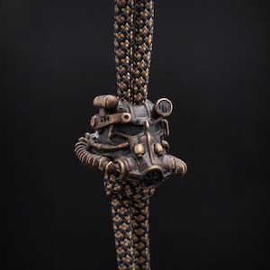 May include: A bronze-colored pendant featuring a detailed replica of a power armor helmet, suspended by a black and brown braided cord. The helmet has intricate mechanical details and a weathered appearance, set against a dark background.