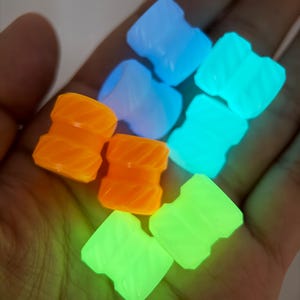 Puede incluir: Una colección de tapones brillantes en varios colores, incluyendo naranja, azul y verde. Los tapones tienen una superficie texturizada y se sostienen en una mano. Los tapones son pequeños y parecen estar hechos de un material translúcido.