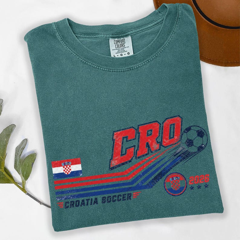 Puede incluir: Una camiseta de cuello redondo color verde azulado con un gr&aacute;fico desgastado que incluye el texto CRO, un bal&oacute;n de f&uacute;tbol, la bandera de Croacia y el texto CROATIA SOCCER 2028. La prenda tiene una etiqueta Comfort Colors de talla grande.