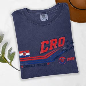 Puede incluir: Una camiseta azul marino de cuello redondo con un gr&aacute;fico que incluye el texto CRO, un bal&oacute;n de f&uacute;tbol, la bandera de Croacia y el texto 2026 y CROATIA SOCCER. La etiqueta de la marca Comfort Colors en el cuello indica una talla grande.