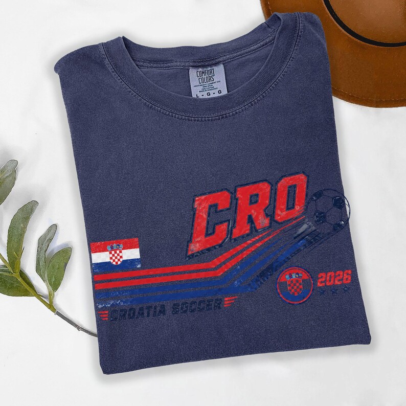 Puede incluir: Una camiseta azul marino de cuello redondo con un gr&aacute;fico que incluye el texto CRO, un bal&oacute;n de f&uacute;tbol, la bandera de Croacia y el texto 2026 y CROATIA SOCCER. La etiqueta de la marca Comfort Colors en el cuello indica una talla grande.