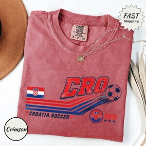 Puede incluir: Una camiseta de manga corta color carmes&iacute; con un gr&aacute;fico que incluye el texto CRO, un bal&oacute;n de f&uacute;tbol, la bandera de Croacia y las palabras CROATIA SOCCER 2023. La prenda tiene una etiqueta Comfort Colors en el cuello y una cadena plateada.