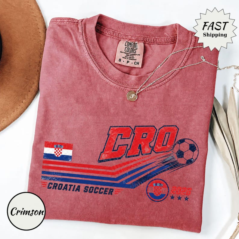 Puede incluir: Una camiseta de manga corta color carmes&iacute; con un gr&aacute;fico que incluye el texto CRO, un bal&oacute;n de f&uacute;tbol, la bandera de Croacia y las palabras CROATIA SOCCER 2023. La prenda tiene una etiqueta Comfort Colors en el cuello y una cadena plateada.
