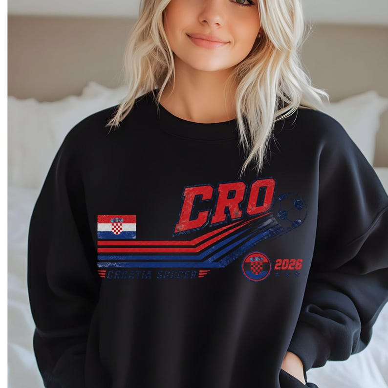 Puede incluir: Una sudadera negra de manga larga con un gr&aacute;fico desgastado que muestra el texto CRO, la bandera de Croacia y las palabras CROATIA SOCCER. El dise&ntilde;o incluye rayas horizontales rojas y azules, un bal&oacute;n de f&uacute;tbol y un emblema con el a&ntilde;o 2026.