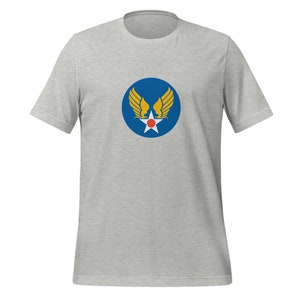May include: A light gray t-shirt featuring a blue circle with a white star, red dot, and yellow wings design. The shirt has short sleeves and a crew neck. The graphic is centered on the chest.