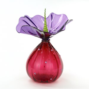 May include: A decorative glass sculpture in the shape of a flower. The sculpture features a deep red, teardrop-shaped base adorned with small, clear accents. A large, translucent purple petal-like structure flares out from the top, with a green, spiral accent in the center.