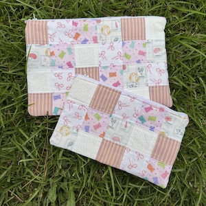 Peut inclure: Deux pochettes rectangulaires à fermeture éclair avec un motif patchwork en tissu matelassé. Le design alterne des carrés de tissu rose et blanc avec de petits motifs d'oursons en gomme, des rubans roses et des carrés à rayures beiges.