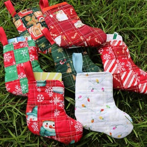 May include: A collection of small, quilted fabric Christmas stockings featuring various holiday patterns including gingerbread men, snowflakes, candy canes, snowmen, and colorful string lights, each equipped with a fabric hanging loop at the top.