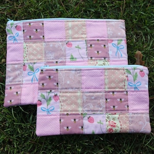 Peut inclure: Deux pochettes rectangulaires zippées en tissu matelassé avec un motif patchwork. Le design comprend des carrés ornés d'abeilles, de fraises, de nœuds bleus et de motifs floraux dans des tons rose, blanc et vert clair avec des fermetures éclair bleu clair.
