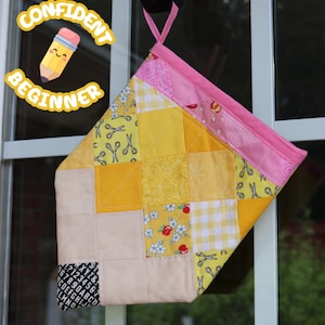 Op de afbeelding: Een gewatteerde pannenlap met een patchworkontwerp van gele, crèmekleurige en roze stoffen met patronen van scharen, bloemen en vichyruitjes. Het heeft een roze stoffen lus om op te hangen en een zwarte hoek met witte letters en cijfers.