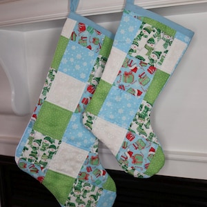 May include: Two patchwork Christmas stockings featuring a grid pattern of light blue, lime green, and white fabric squares. The fabric prints include snowmen, wrapped gifts, and snowflakes. Each stocking has a light blue trim and a hanging loop.