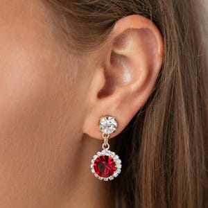 May include: Gold dangle earrings with a red crystal surrounded by smaller clear crystals and a smaller clear crystal at the top.