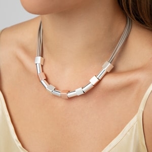 May include: A silver and rose gold necklace with a layered leather cord and square-shaped beads.