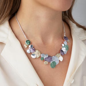 Puede incluir: Un collar plateado con una serie de colgantes circulares de colores superpuestos. Los colgantes son en tonos verdes, morados, plateados y dorados. El collar se lleva sobre una camisa blanca.
