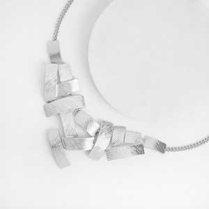 May include: Silver-toned necklace with a geometric design. The necklace features rectangular, textured metal pieces arranged in an asymmetrical pattern. The chain is a delicate, linked design. The background is white.