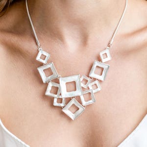 Peut inclure: Un collier argenté avec un design géométrique composé de plusieurs carrés blancs de tailles différentes.