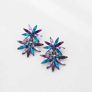 May include: A pair of dangle earrings featuring a purple and blue flower design. The earrings are made of metal and have a silver finish.