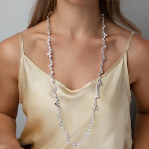 Op de afbeelding: Een lange, zilveren ketting met een delicaat ontwerp van gegroepeerde, sprankelende stenen. De ketting wordt gedragen tegen een lichtgouden satijnen top. De stenen lijken helder en reflecterend. De ketting is ongeveer 70 cm lang.