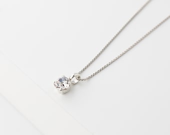 May include: A silver necklace with a single, clear, round-cut gemstone pendant. The pendant is set in a silver bezel and hangs from a delicate silver chain. The background is a soft white.