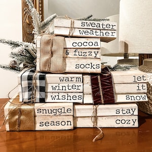 May include: A stack of decorative wooden blocks painted white, with black text and tied with ribbons and twine. The blocks feature phrases like "sweater weather," "cocoa & fuzzy," and "snuggle season." A cozy winter theme.