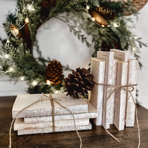 May include: A stack of decorative books tied with twine, featuring aged paper pages. A pine cone rests on top of the books. A Christmas wreath with pinecones and lights is in the background. The books are on a dark wooden surface.