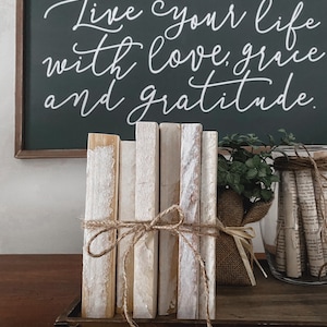 May include: A decorative wooden sign with the text "Live your life with love, grace, and gratitude" in white script. In front of the sign are several white-painted wooden blocks tied with twine, and a small plant.