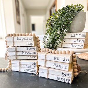 May include: Stacked decorative books with inspirational phrases like "bloom with grace" and "He is Risen." The books are tied with twine and adorned with wooden beads. A potted plant sits in the background.
