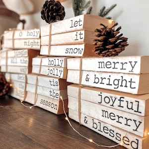 May include: Stacked decorative books with Christmas-themed phrases like "Merry & Bright" and "Let it Snow." The books are tied with twine and adorned with pinecones and string lights, creating a festive holiday display.