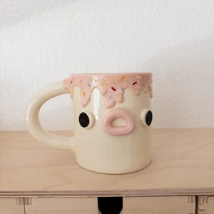 May include: A whimsical ceramic mug with a face design. The off-white mug features black eyes, a pink mouth, and a pink glaze with colorful sprinkles around the rim. The mug has a curved handle.