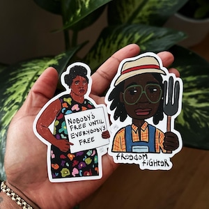 May include: Two stickers featuring illustrations of Black people. The first sticker shows a woman holding a sign that reads "Nobody's Free Until Everybody's Free". The second sticker shows a person wearing a straw hat and overalls holding a pitchfork. The text "Freedom Fighter" is written below the person.