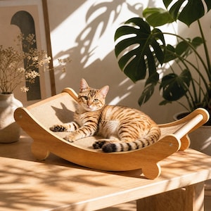 Könnte beinhalten: Eine getigerte Katze liegt auf einer Katzenhängematte aus Holz. Die Hängematte hat ein beiges Stoffbett und einen gebogenen Holzrahmen. Die Katze hat braune und schwarze Streifen. Die Hängematte steht auf einem Holztisch.