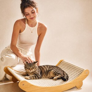 K&ouml;nnte beinhalten: Eine braun-schwarz gestreifte Katze liegt auf einem Katzenbett aus Holz mit einer gewebten Sisaloberfl&auml;che. Das Bett hat ein geschwungenes Design und wird von Holzbeinen getragen. Eine Person streichelt sanft die Katze.