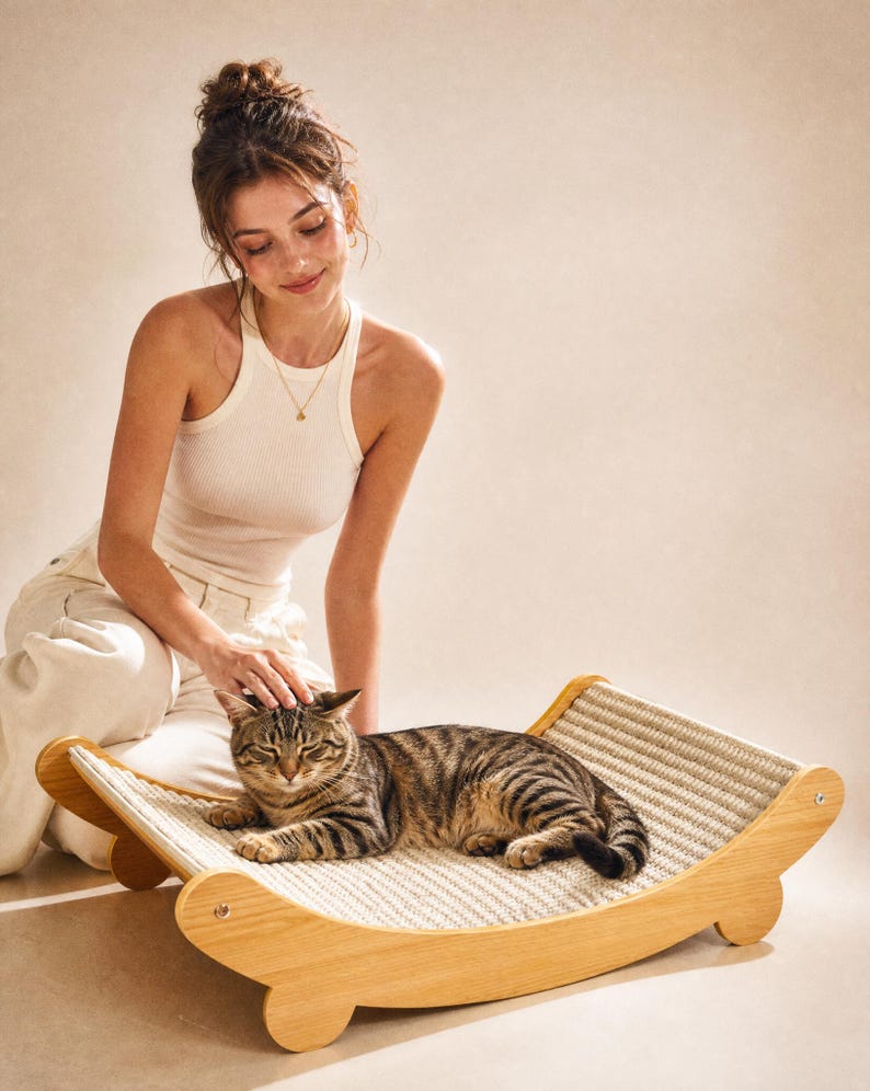 K&ouml;nnte beinhalten: Eine braun-schwarz gestreifte Katze liegt auf einem Katzenbett aus Holz mit einer gewebten Sisaloberfl&auml;che. Das Bett hat ein geschwungenes Design und wird von Holzbeinen getragen. Eine Person streichelt sanft die Katze.