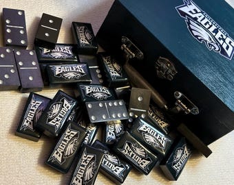 May include: A set of dominoes and a wooden box, featuring the Philadelphia Eagles logo. The dominoes are black with white dots, and the box is dark blue with the team's logo on the lid. A fun game for Eagles fans.