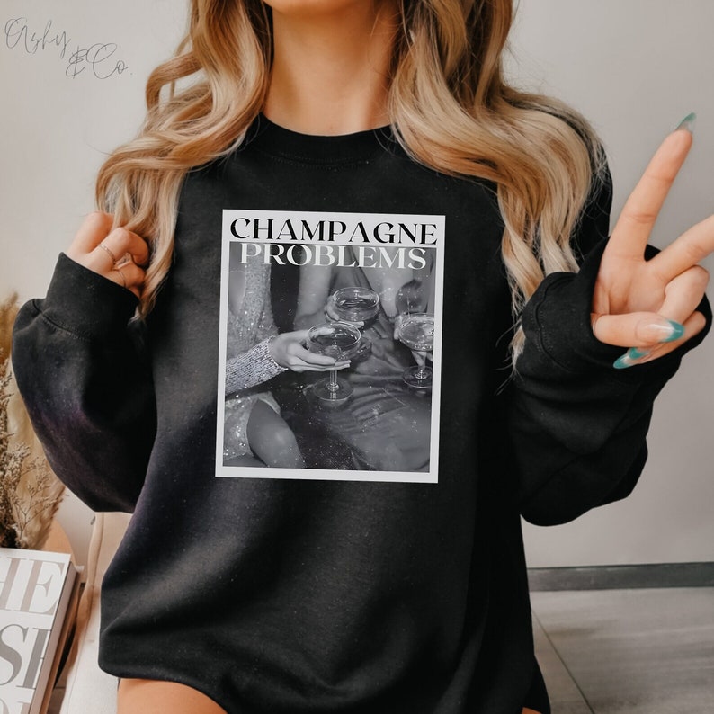May include: Black sweatshirt with a graphic print of three people holding champagne glasses. The text "Champagne Problems" is printed above the image.