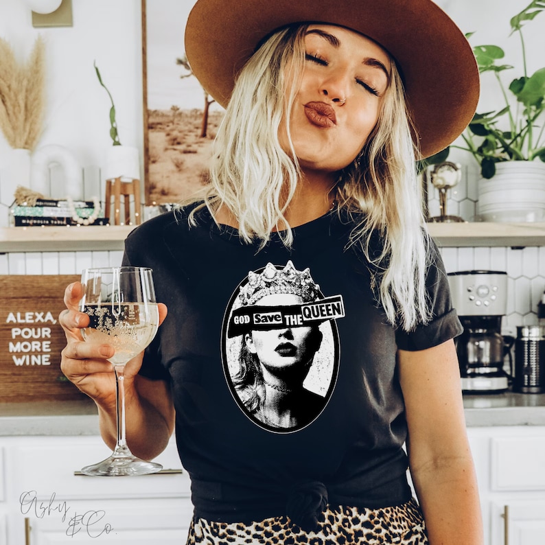 May include: A black t-shirt with a graphic of a woman wearing a crown. The graphic has the text "God Save The Queen" with the word "Queen" partially obscured by a black and white stripe.