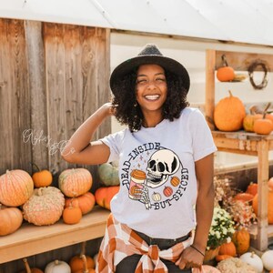 Peut inclure: Un t-shirt blanc avec un graphique d'un squelette tenant une tasse de café et le texte "Dead inside but spiced". Le t-shirt est porté par une personne aux cheveux foncés et un chapeau noir. La personne est debout devant un mur de citrouilles.