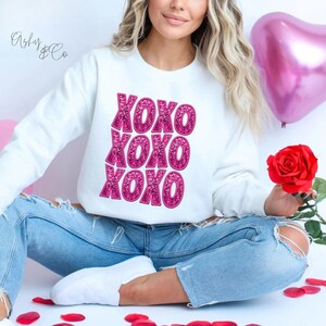 May include: White sweatshirt with pink glitter XOXO text. The sweatshirt is being worn by a person sitting with a rose and a pink heart balloon in the background.