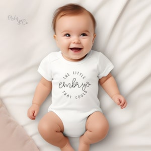 May include: A white baby grow with a black circle design that says "The Little Embryo That Could". A baby is wearing the grow and smiling.