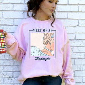 May include: Pink sweatshirt featuring a graphic of Cinderella's silhouette and the text "Meet me at Midnight".