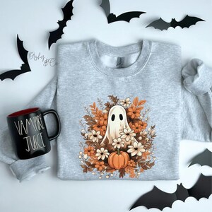 May include: A grey sweatshirt featuring a white ghost surrounded by orange and white flowers and a small orange pumpkin. The ghost has two small eyes and a small mouth.