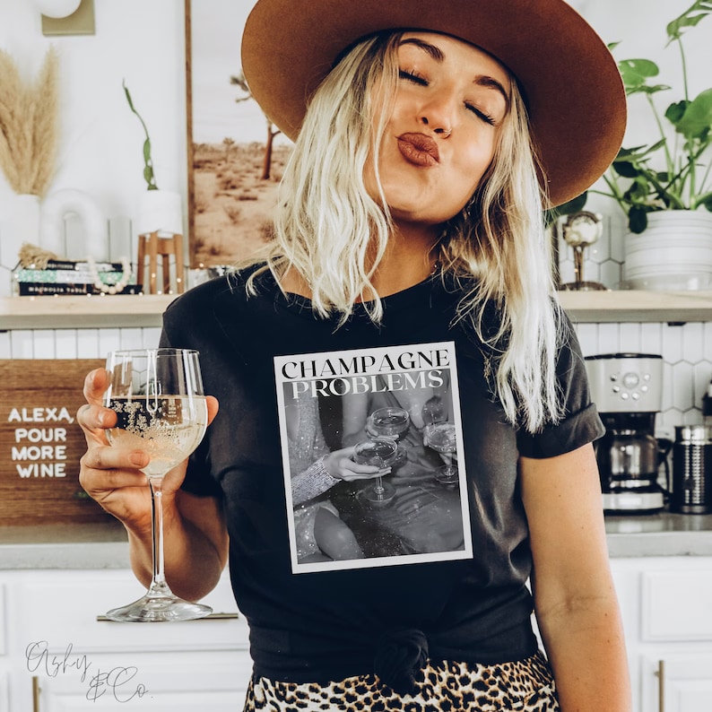 May include: A black t-shirt with the text "Champagne Problems" printed on the front. The text is in white and is surrounded by a black and white image of people holding champagne glasses.