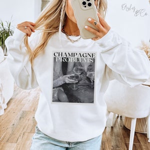May include: White sweatshirt with a black and white graphic print of three people holding champagne glasses. The text "Champagne Problems" is printed above the graphic.