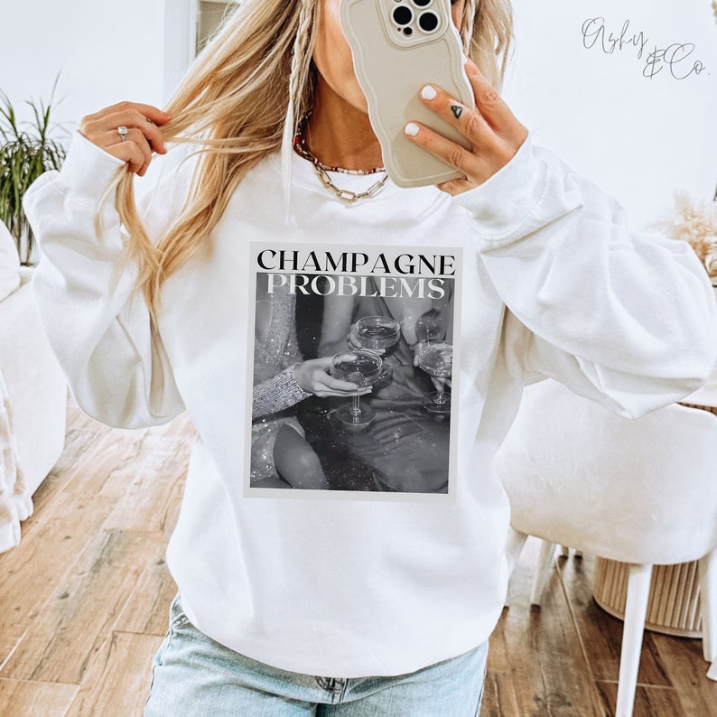 May include: White sweatshirt with a black and white graphic print of three people holding champagne glasses. The text "Champagne Problems" is printed above the graphic.