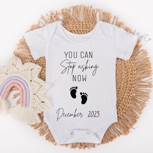 May include: White baby onesie with black text that reads "You can stop asking now December 2023".  Two black footprints are above the date.