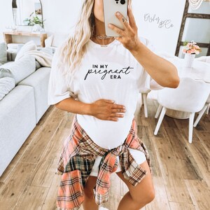 May include: White t-shirt with the text "In My Pregnant Era" in black script font. The person wearing the shirt is holding their stomach.
