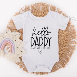 May include: White baby onesie with black text that reads "hello daddy I can't wait to meet you" with a small heart symbol.