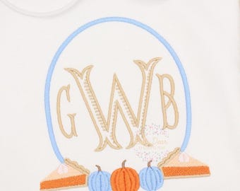 Op de afbeelding: Wit geborduurd monogram met de letters GWB in een lichtblauwe cirkel, omringd door drie oranje pompoenen en twee blauwe pompoenen, en twee oranje taartbodems.